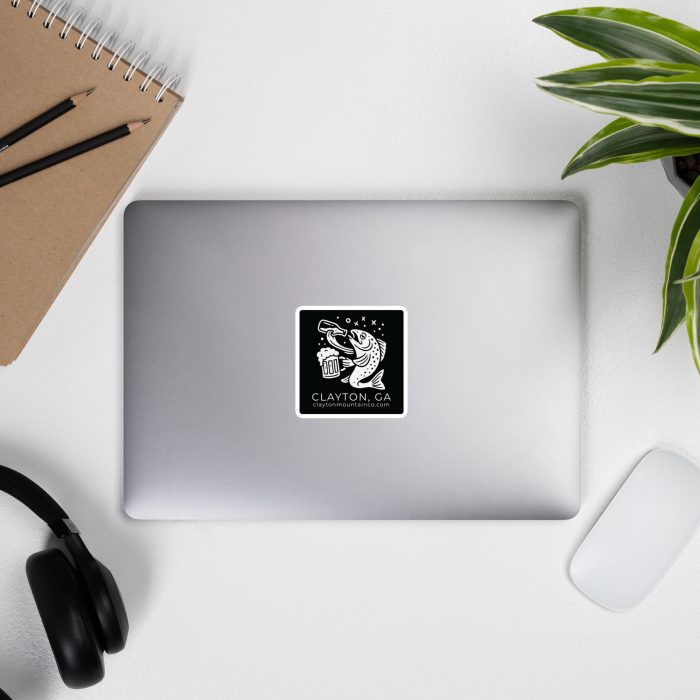 die-cut-stickers-satin-3x3-front-6a2e9f73368e4jpg - Clayton Mountain Co. Top-down view of a desk with a spiral notebook, two pencils, black headphones, a closed silver laptop, a green plant, and a white wireless mouse on a white surface.